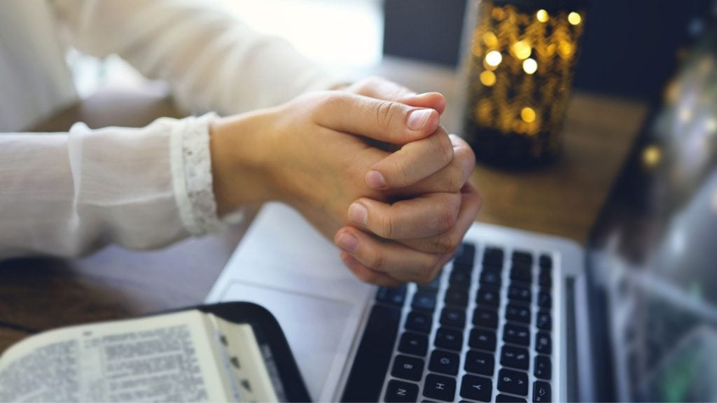 christian-blogger-sitting-at-her-desk-with-hand-together-over-a-laptop-and-next-to-a-bible