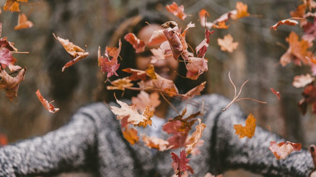 woman in a sweater tossing a pile of autumn leaves in the air